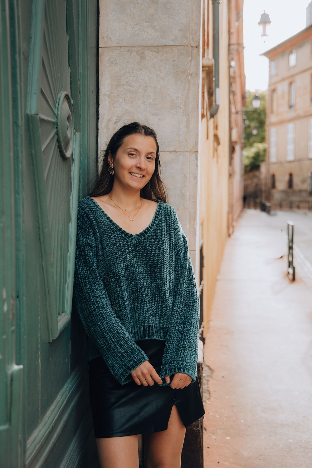 A smiling young woman in a teal knit sweater and black skirt leans against a wall in a European alley.
