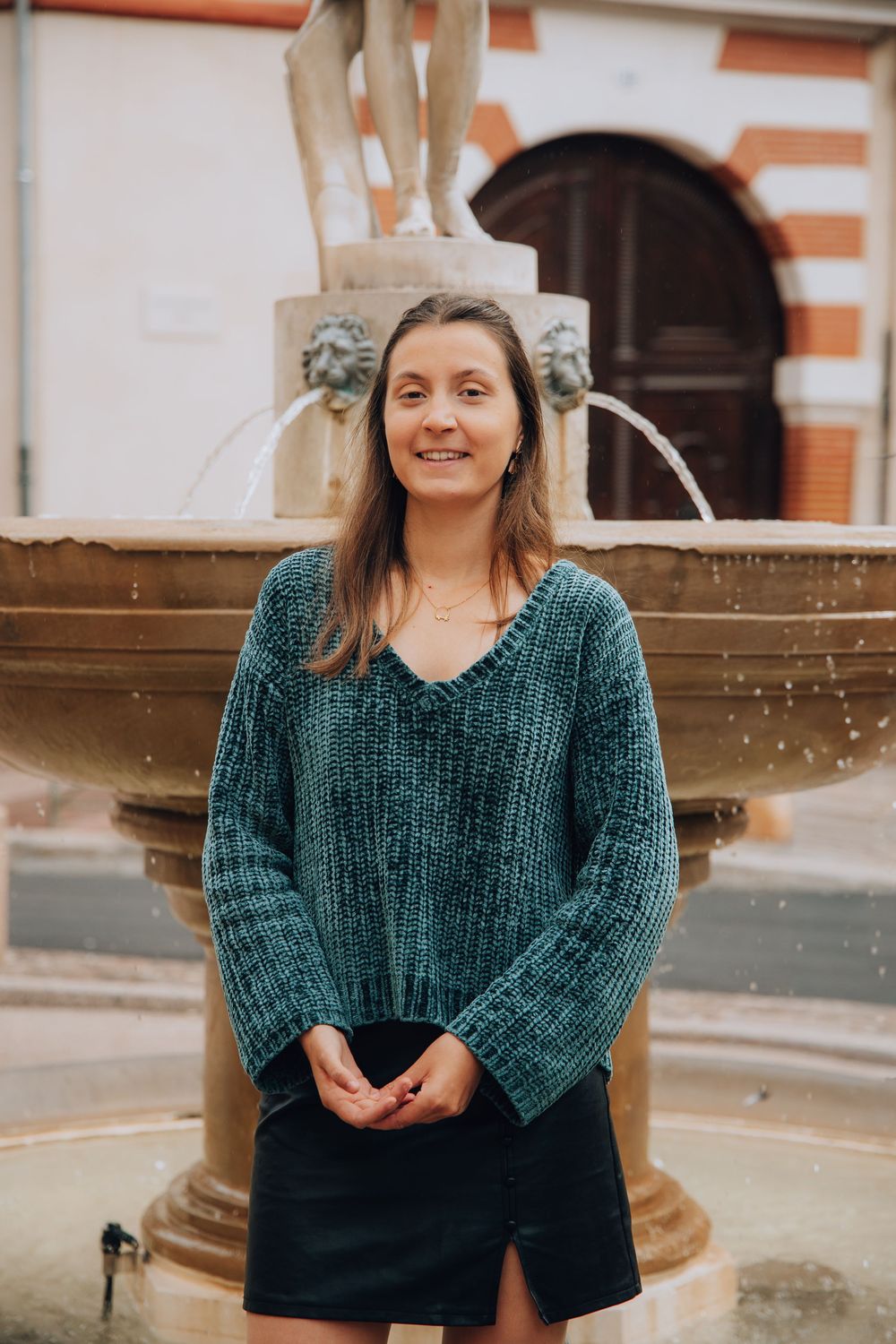 A smiling woman in a teal knit sweater stands in front of an ornate stone fountain in a European square.