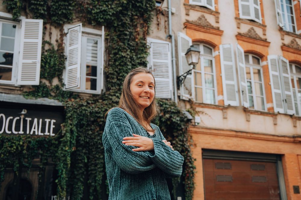 A smiling woman in a teal knit sweater stands with arms crossed outside a building covered in ivy.