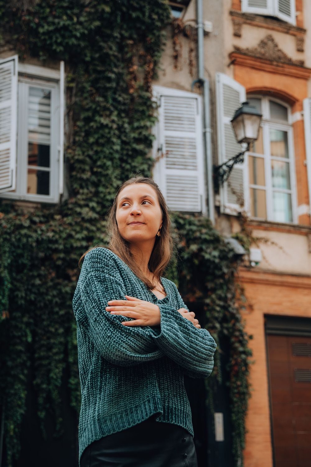 A woman in a teal knit sweater stands with arms crossed, gazing upward in front of an ivy-covered building.