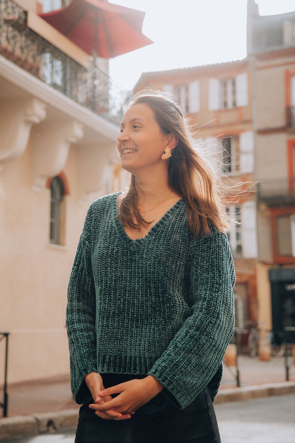 A smiling woman in a teal knit sweater with floral earrings stands on a sunny European street.