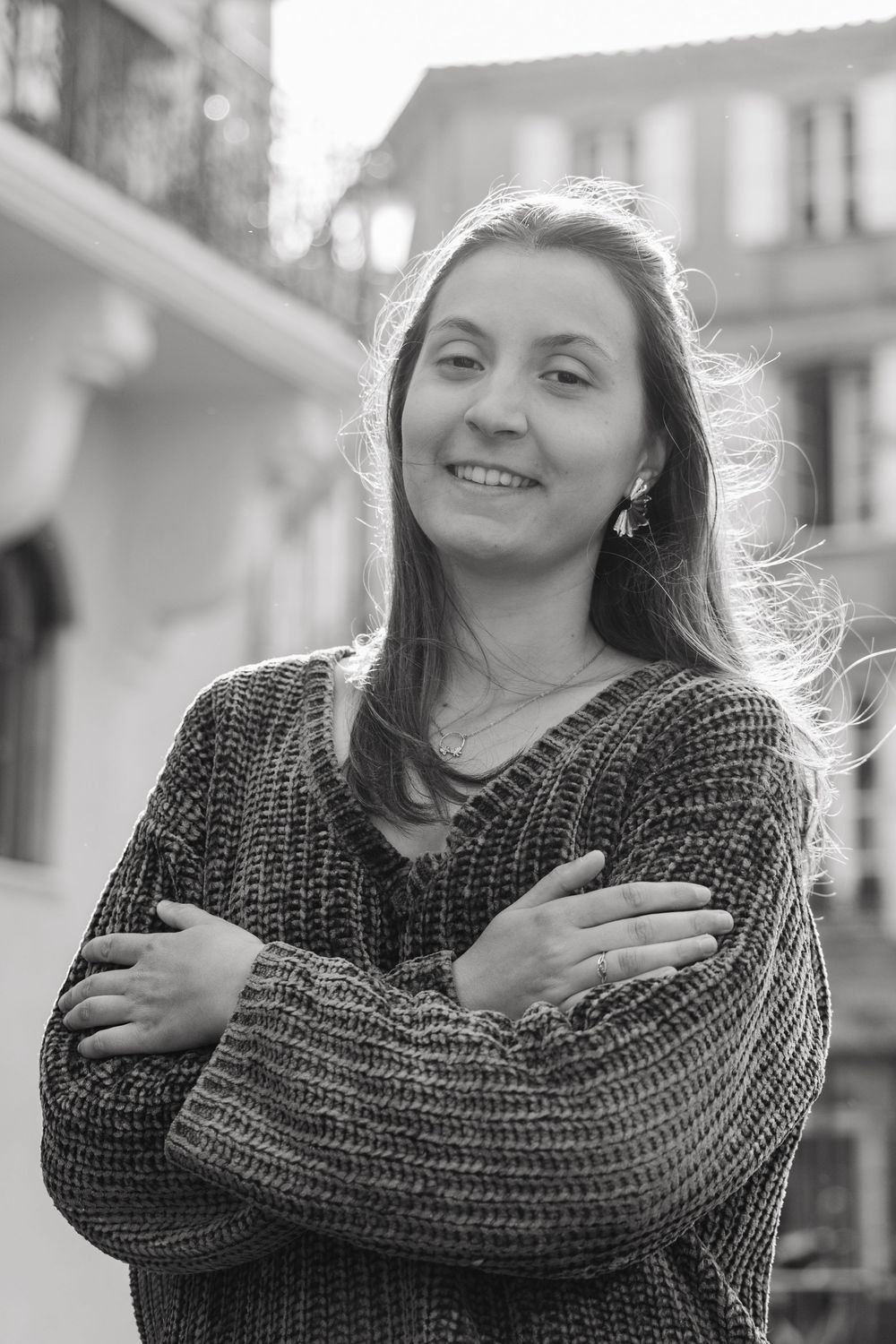 Black and white portrait of a smiling woman in a knit sweater with arms crossed on a European street.