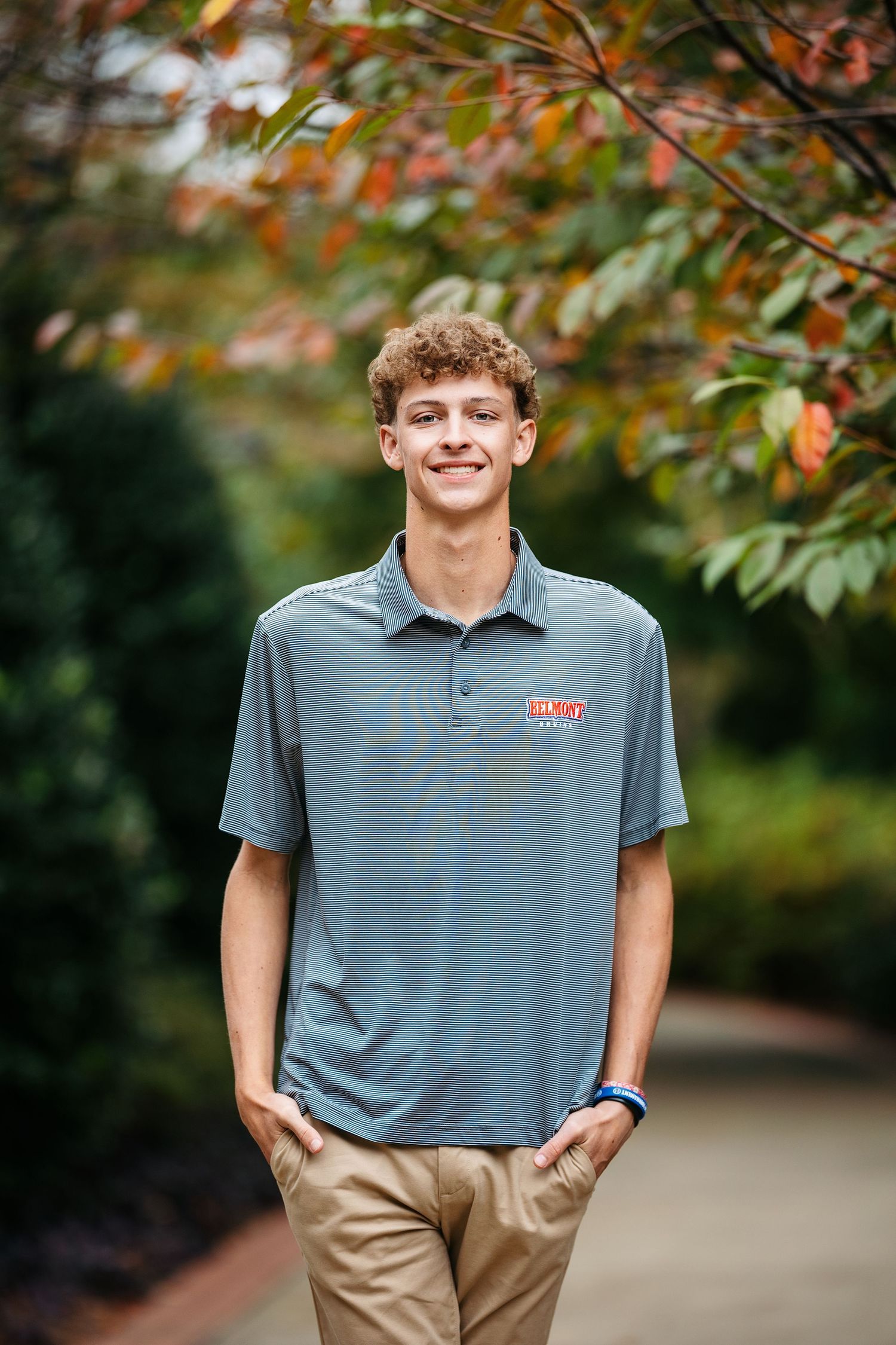 Person in navy shirt and khaki pants poses against white columns of a building exterior with brick walls and trees.