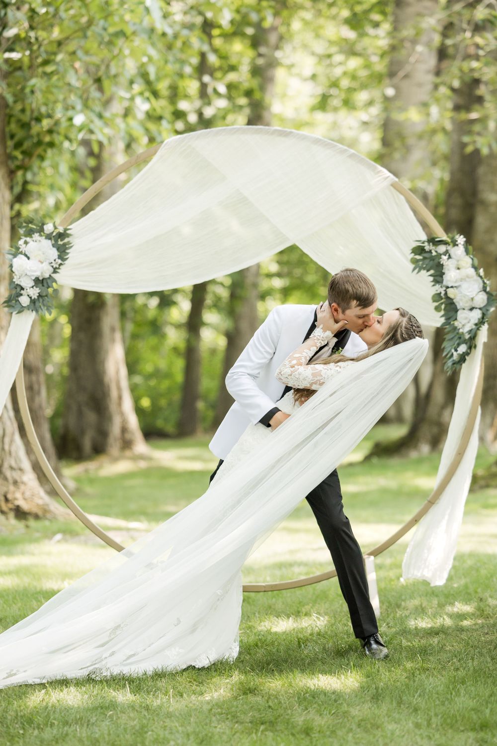 A romantic wedding ceremony setup with flowing white fabric arch and floral decorations in a wooded setting.