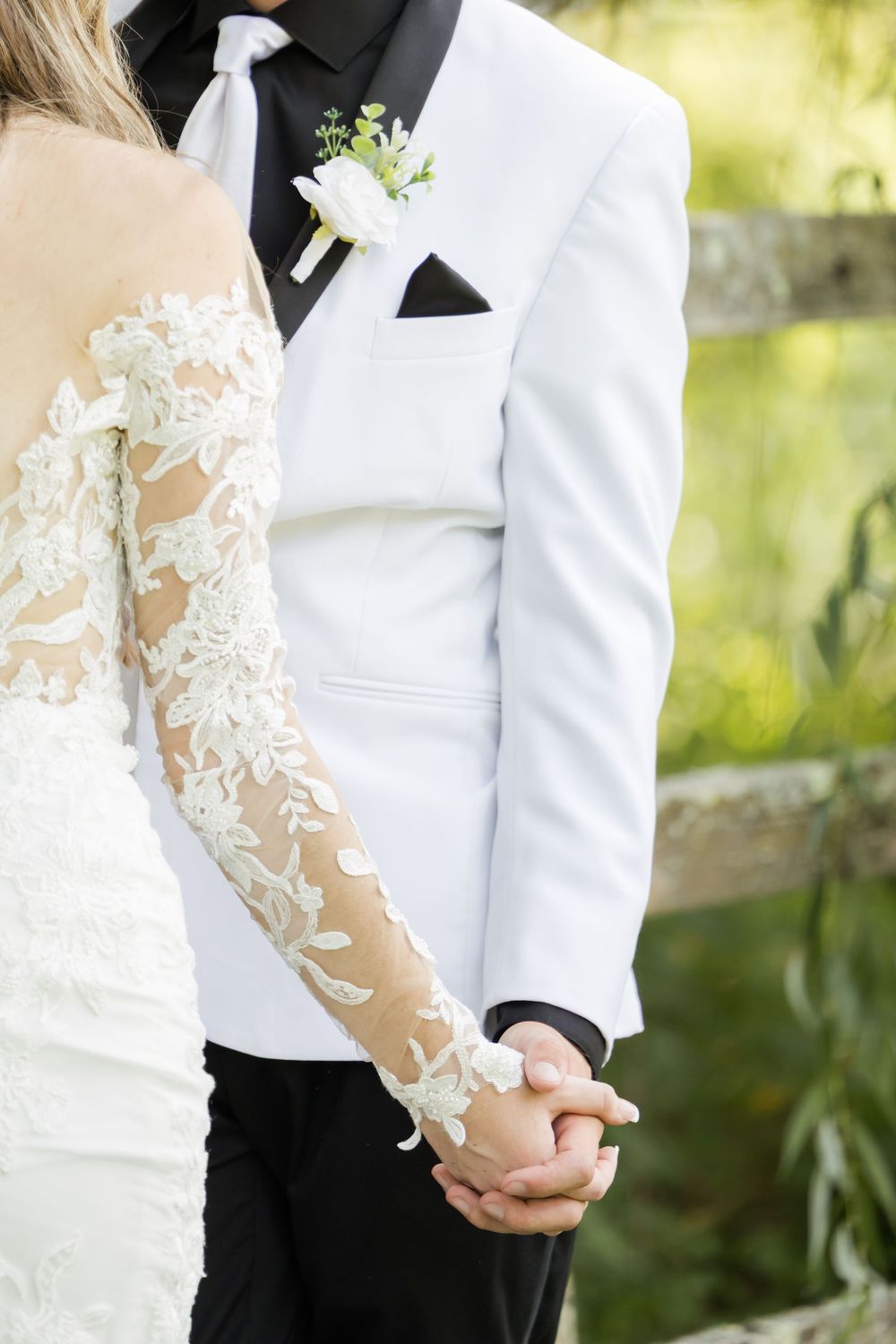 Close-up detail of white lace wedding dress sleeve and groom's white tuxedo jacket with boutonniere.