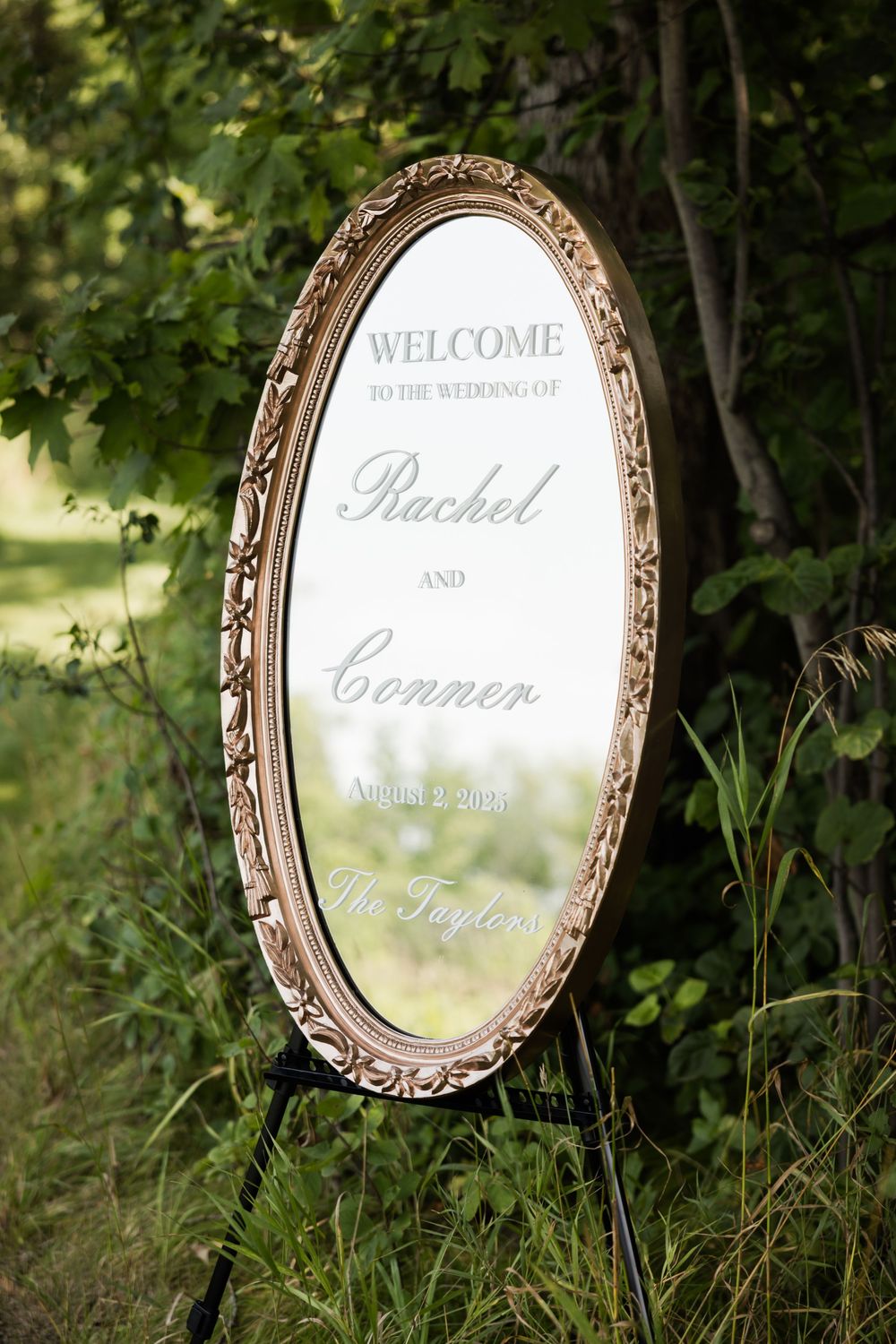 An ornate vintage mirror displays a wedding welcome sign among green foliage.