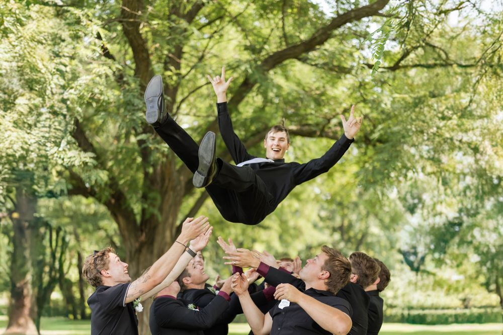 Groomsmen toss a person in formal attire into the air during playful wedding photo moment.