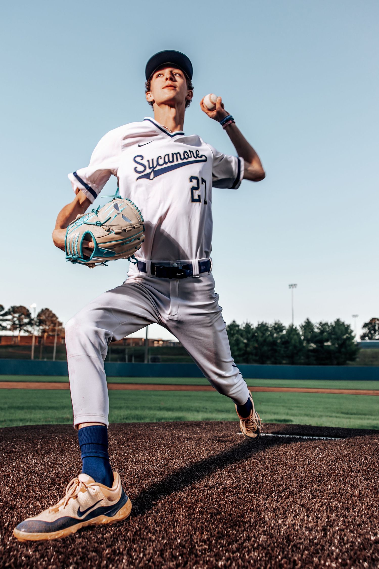 Baseball pitcher in white Sycamore uniform winds up for a throw from the mound during daytime game.