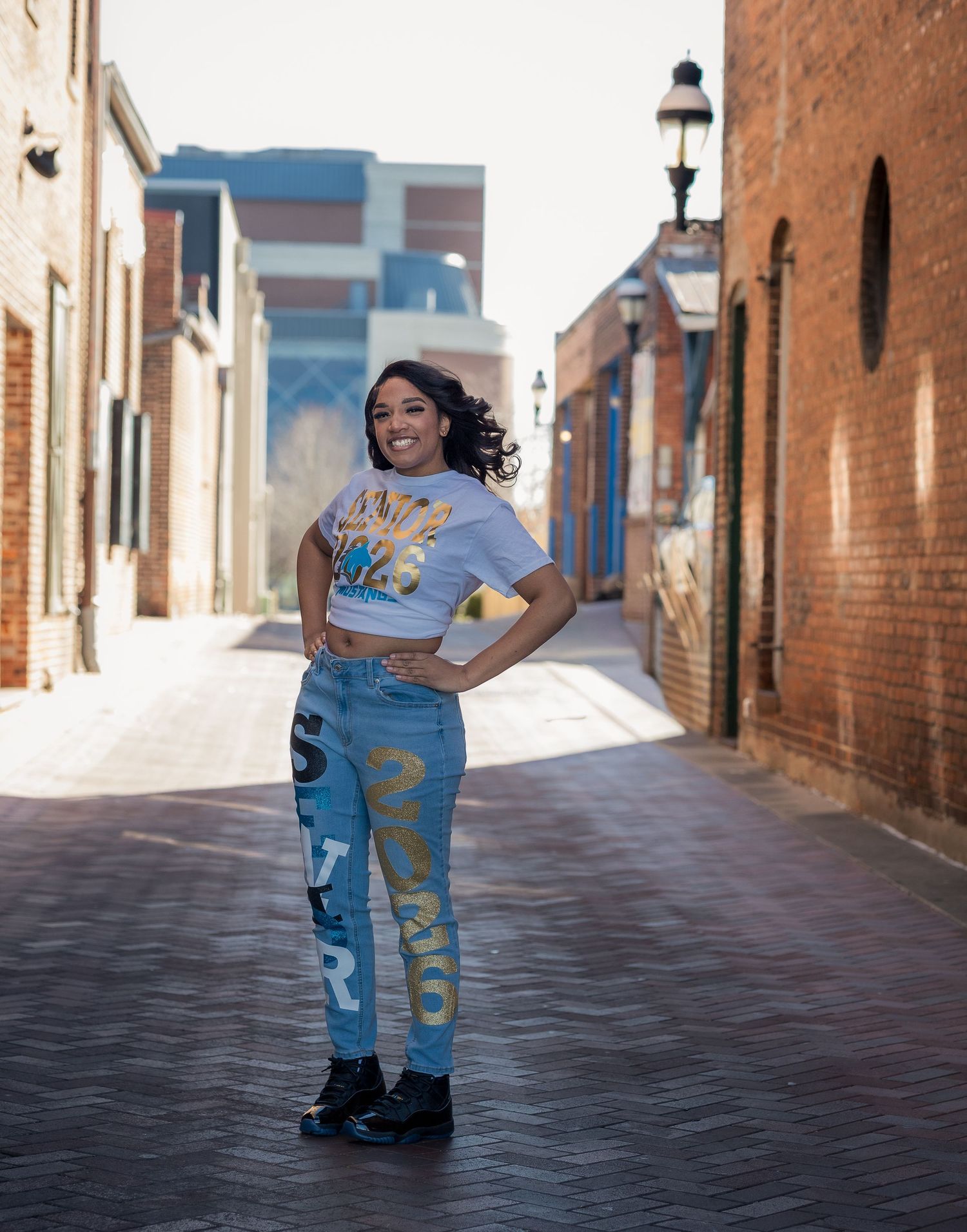 A person in a light blue t-shirt and patterned jeans poses confidently in a brick-lined alley.