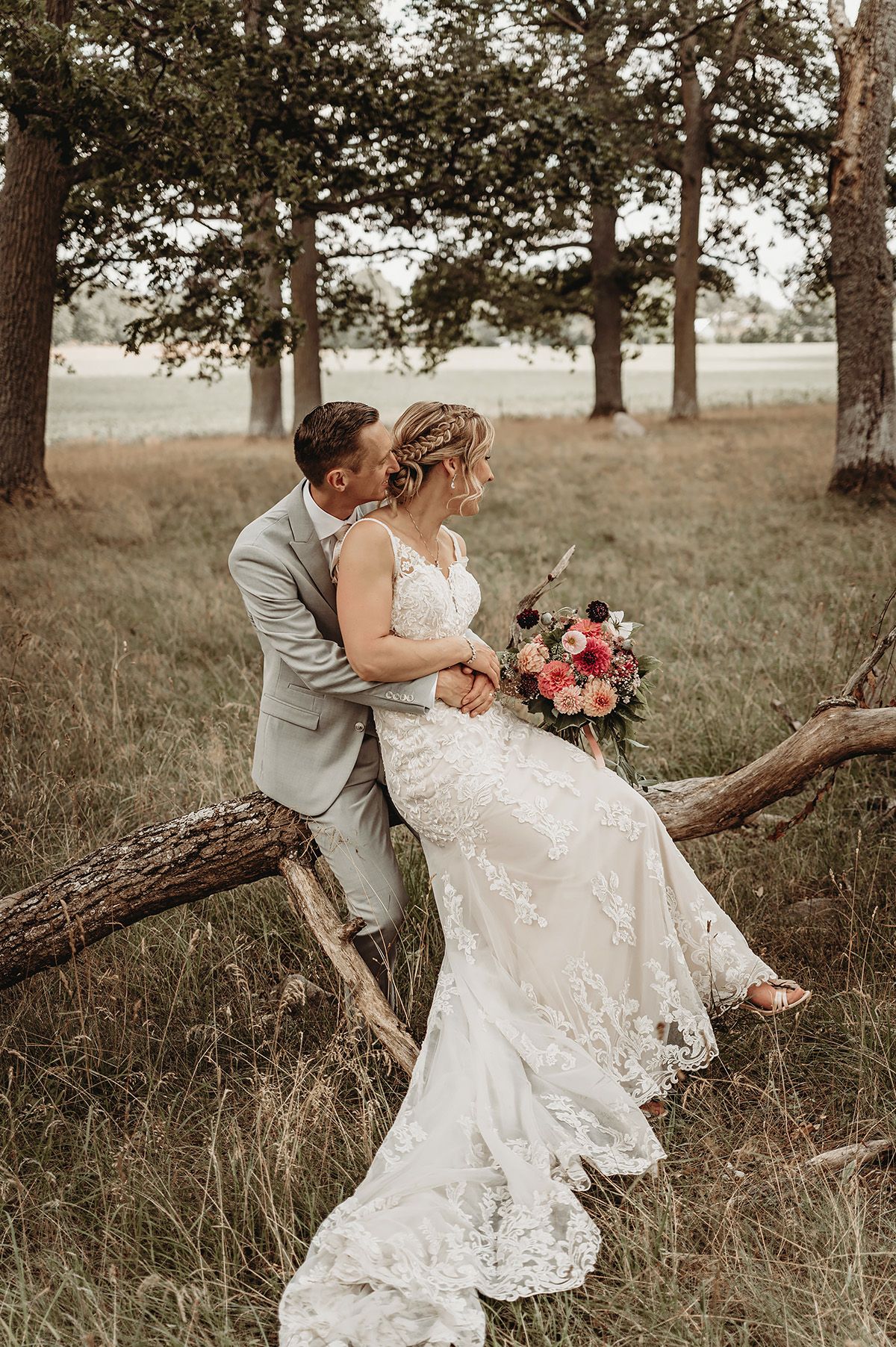 Bröllopsporträtt i grönskande hage med höga ekar i Borensberg, brudparet sitter på ett nedfallet träd. Foto: Lets Bloom Media, Östergötland.