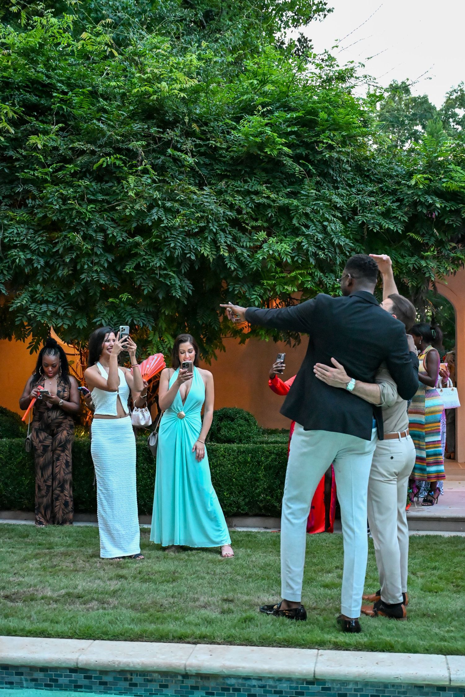 A group of wedding guests in formal attire gather outdoors in a garden setting during a celebration.