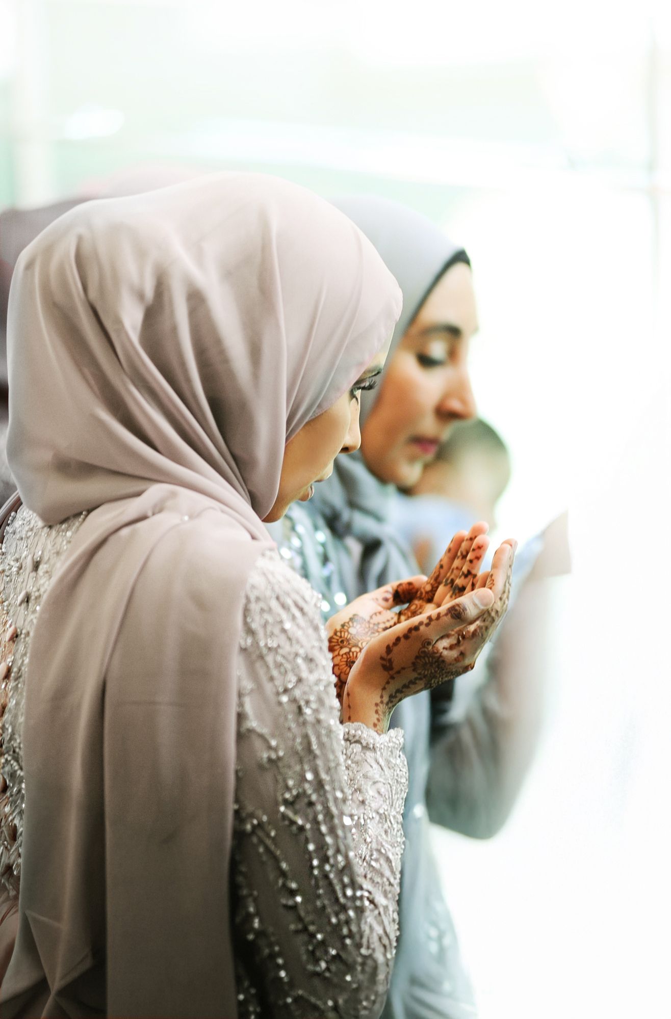 Two Muslim women in hijabs with henna-decorated hands raised in prayer during wedding celebration.