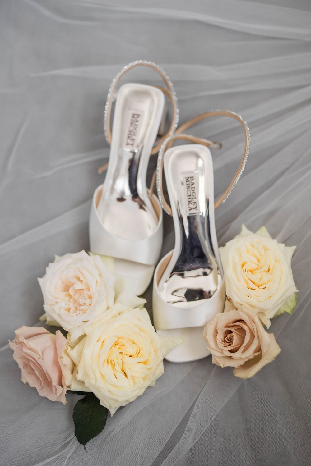 White bridal shoes with delicate peach and cream roses arranged on a soft gray tulle background.