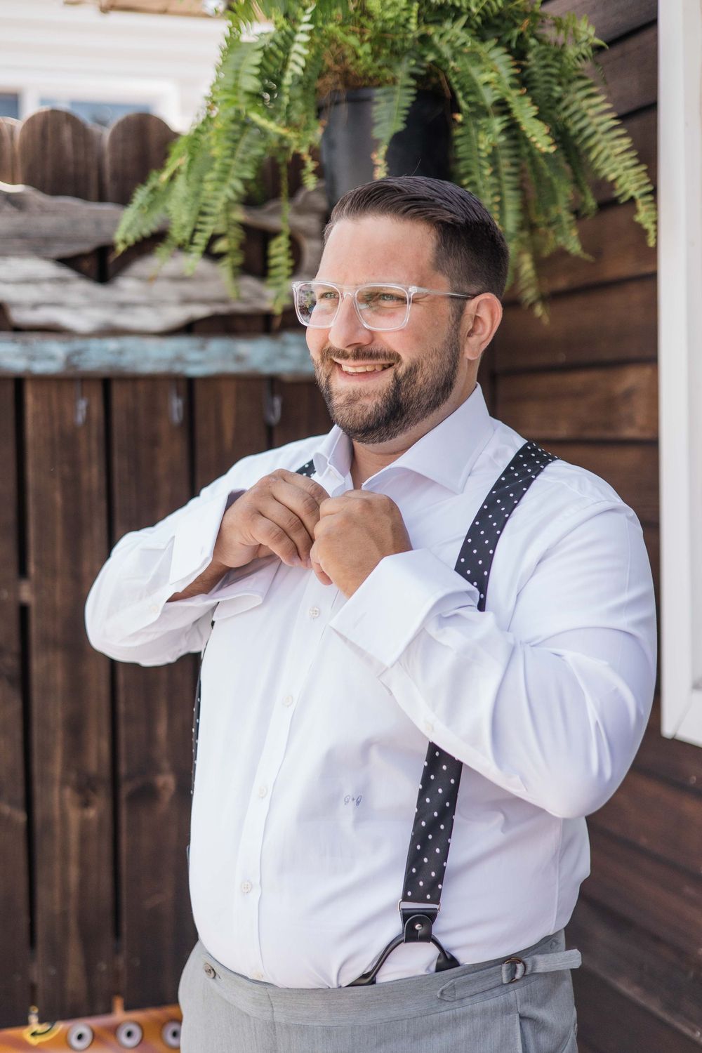 Smiling figure in a crisp white dress shirt and polka dot suspenders adjusts their collar against a wooden backdrop with ferns.