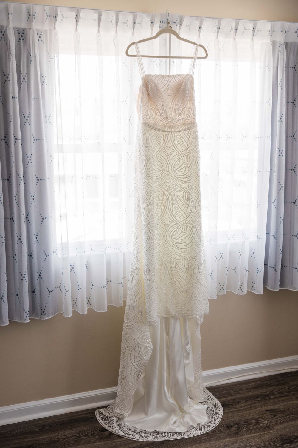 A white lace wedding dress hangs from a hanger against a window with sheer curtains in natural light.