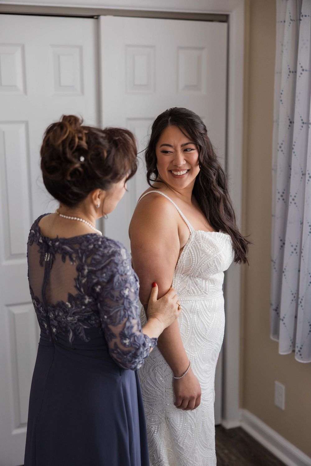 A bride in a white lace dress gets help with final preparations from someone wearing a navy blue dress.