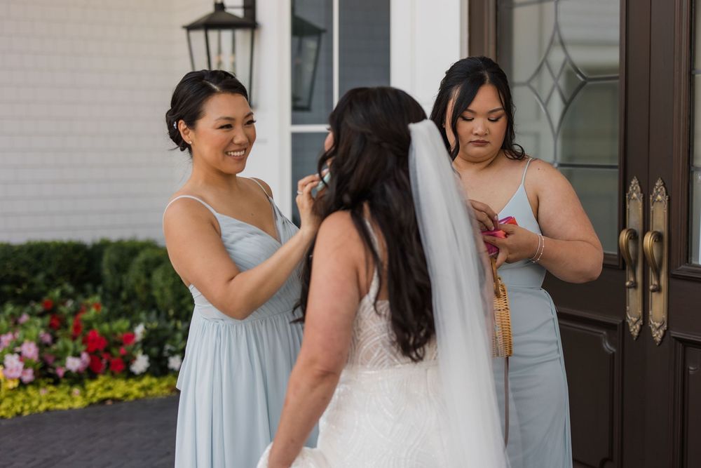 Bridesmaids in light blue dresses help adjust the bride's gown and veil outside a building entrance.