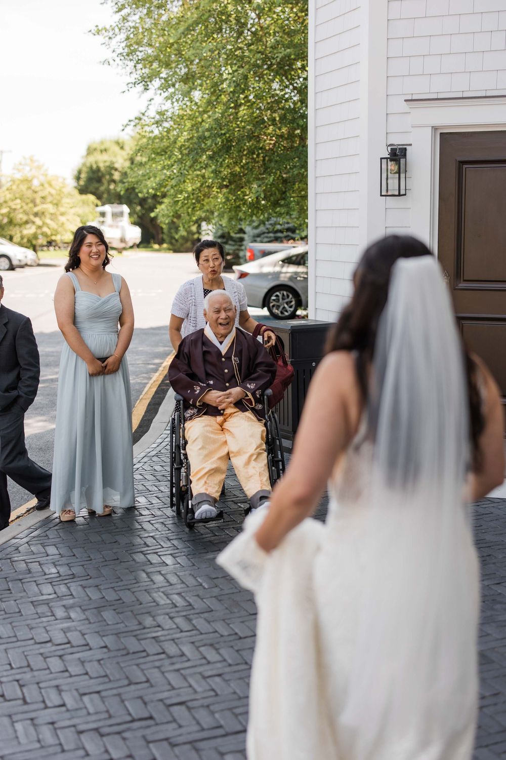 A wedding party gathers on a brick patio outside a white building on a sunny day.