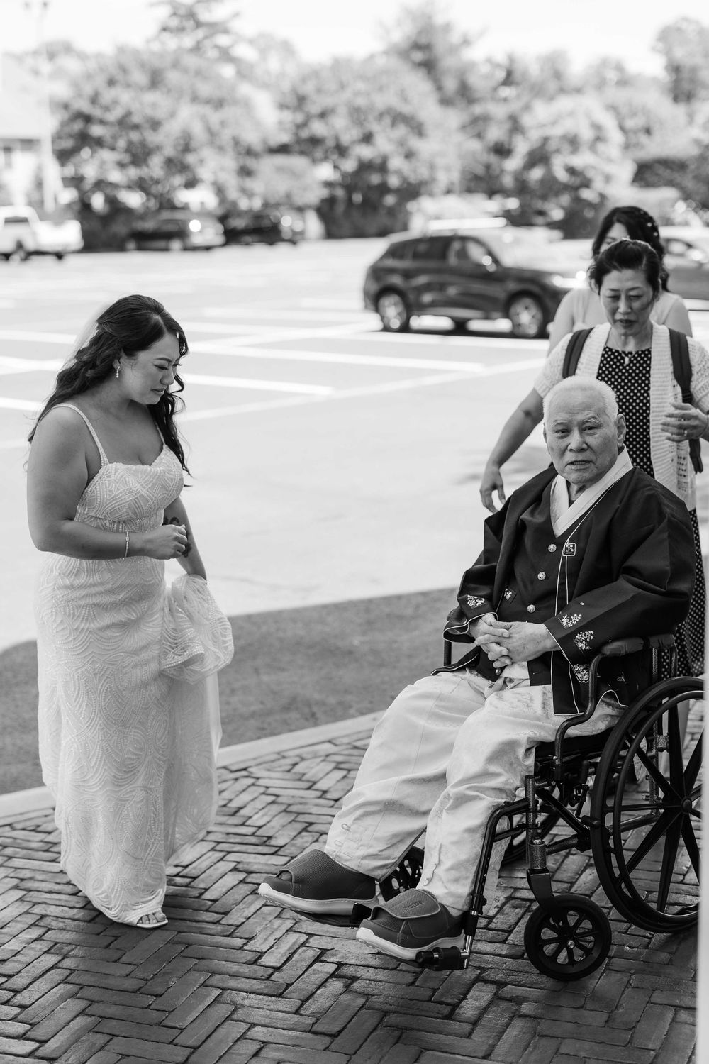 Black and white photo of a bride in a white dress standing next to a person in a wheelchair on a brick pathway.