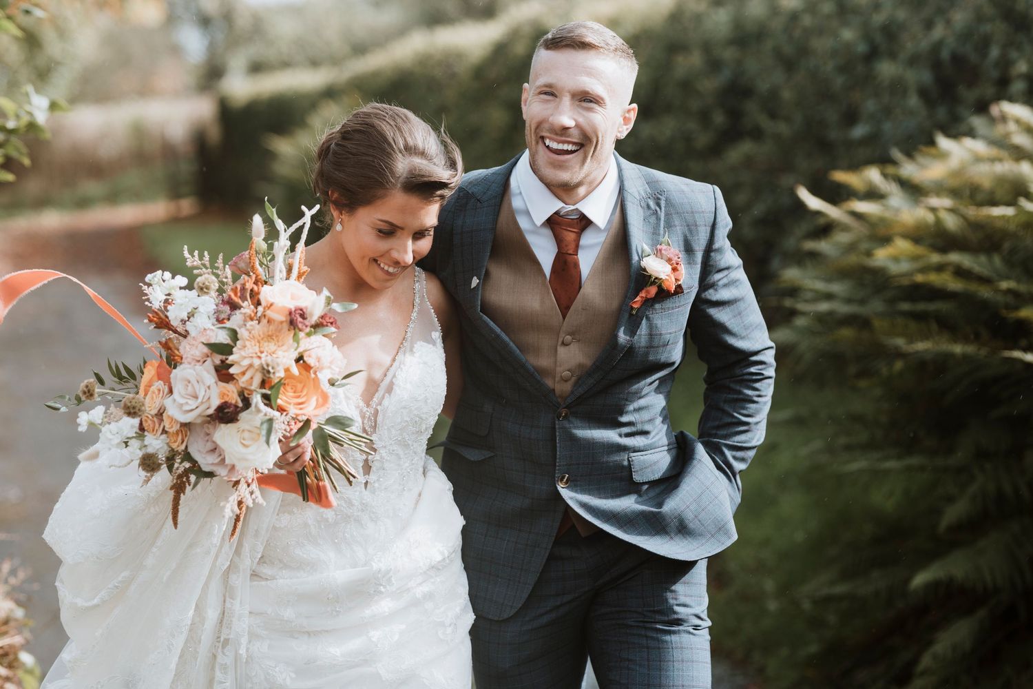 A joyful bride and groom share a candid moment outdoors, with the bride holding a peach and white floral bouquet at Camlad Barns