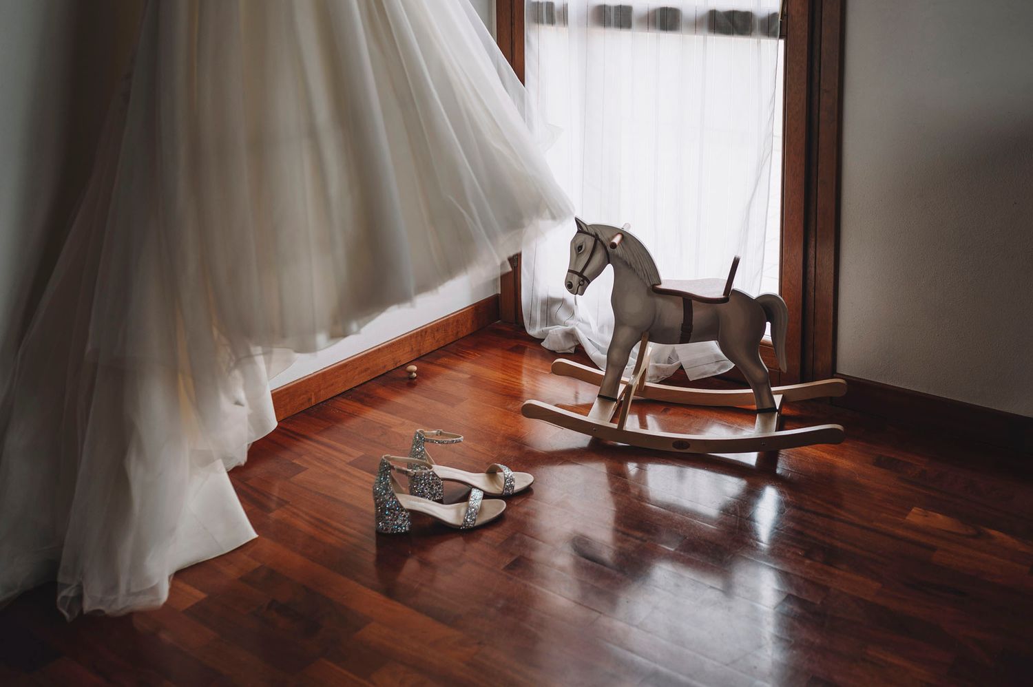 Dettagli di scarpe e abito da sposa disposti su un pavimento in legno massello illuminato dalla morbida luce che entra dalla finestra.