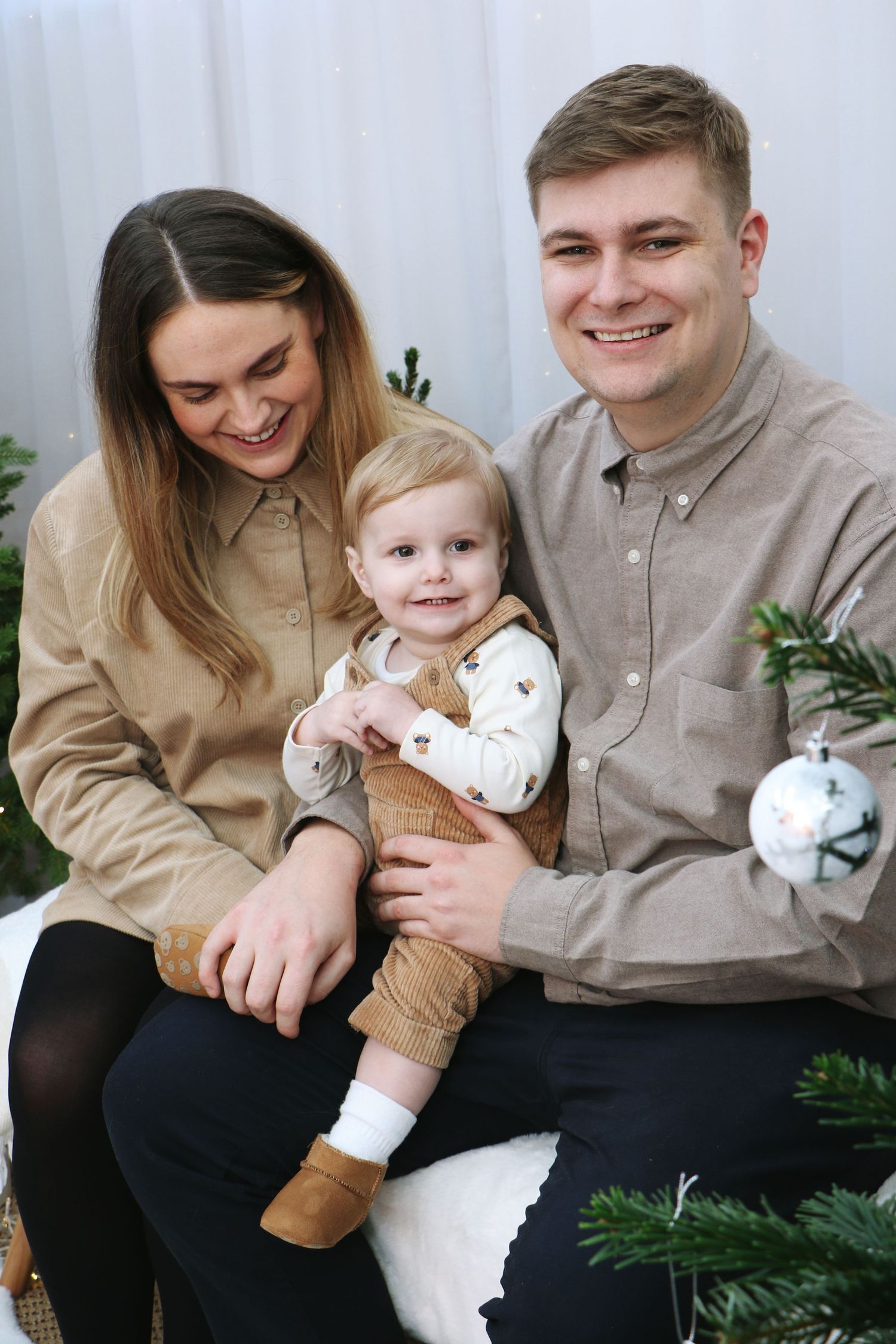 A family poses together for a holiday portrait near a Christmas tree, wearing coordinating beige and brown outfits.