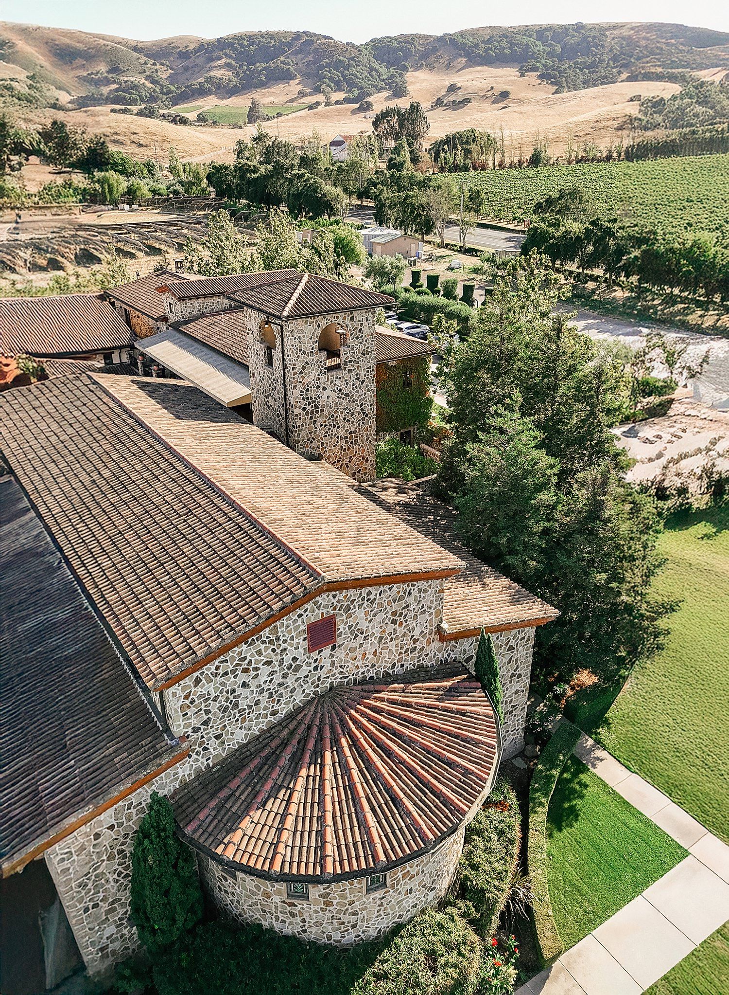 Aerial view of a stone and terracotta winery building with curved architecture nestled in rolling vineyard hills.