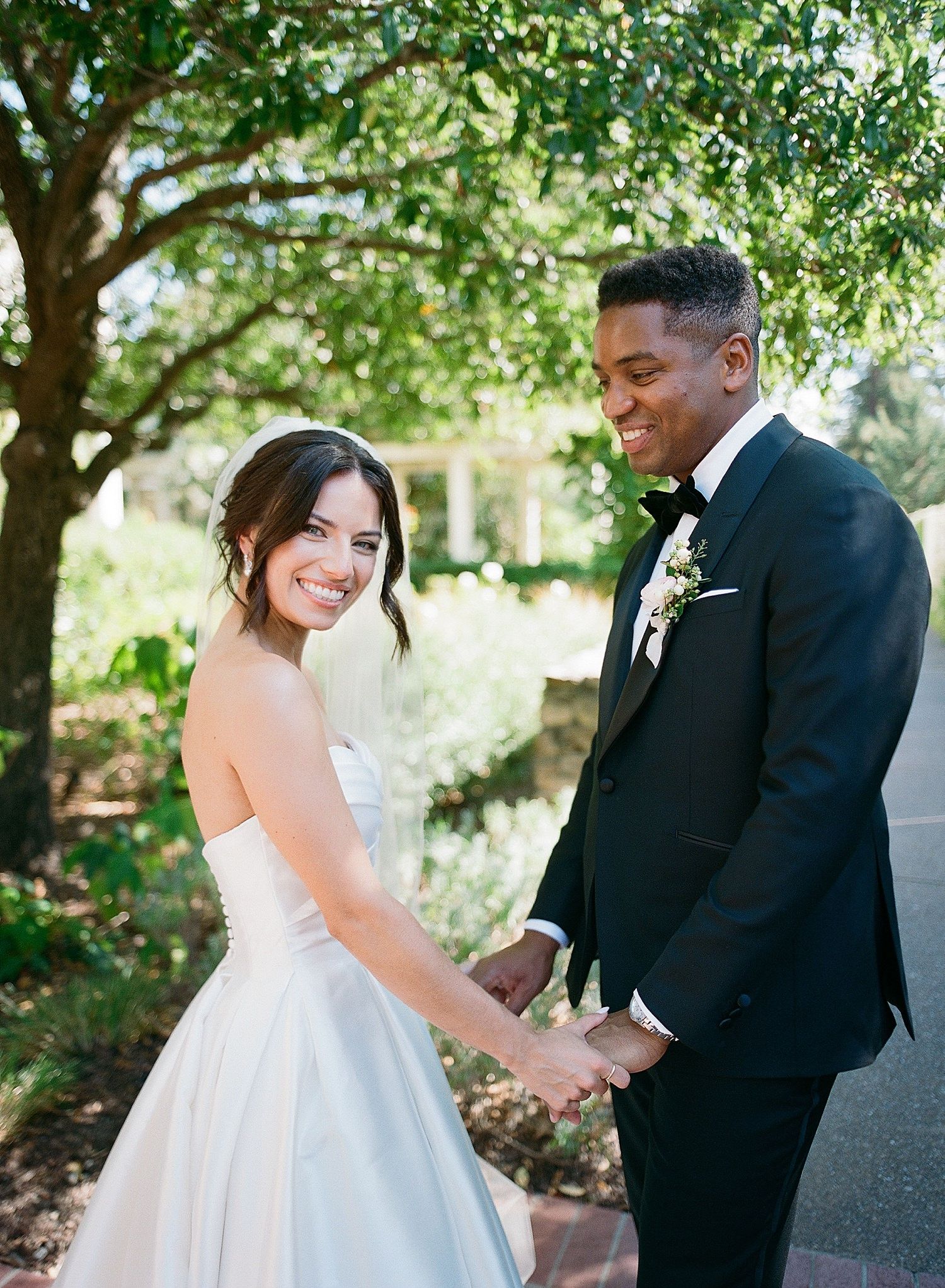 A couple shares a joyful moment together outdoors under green trees during their wedding celebration.