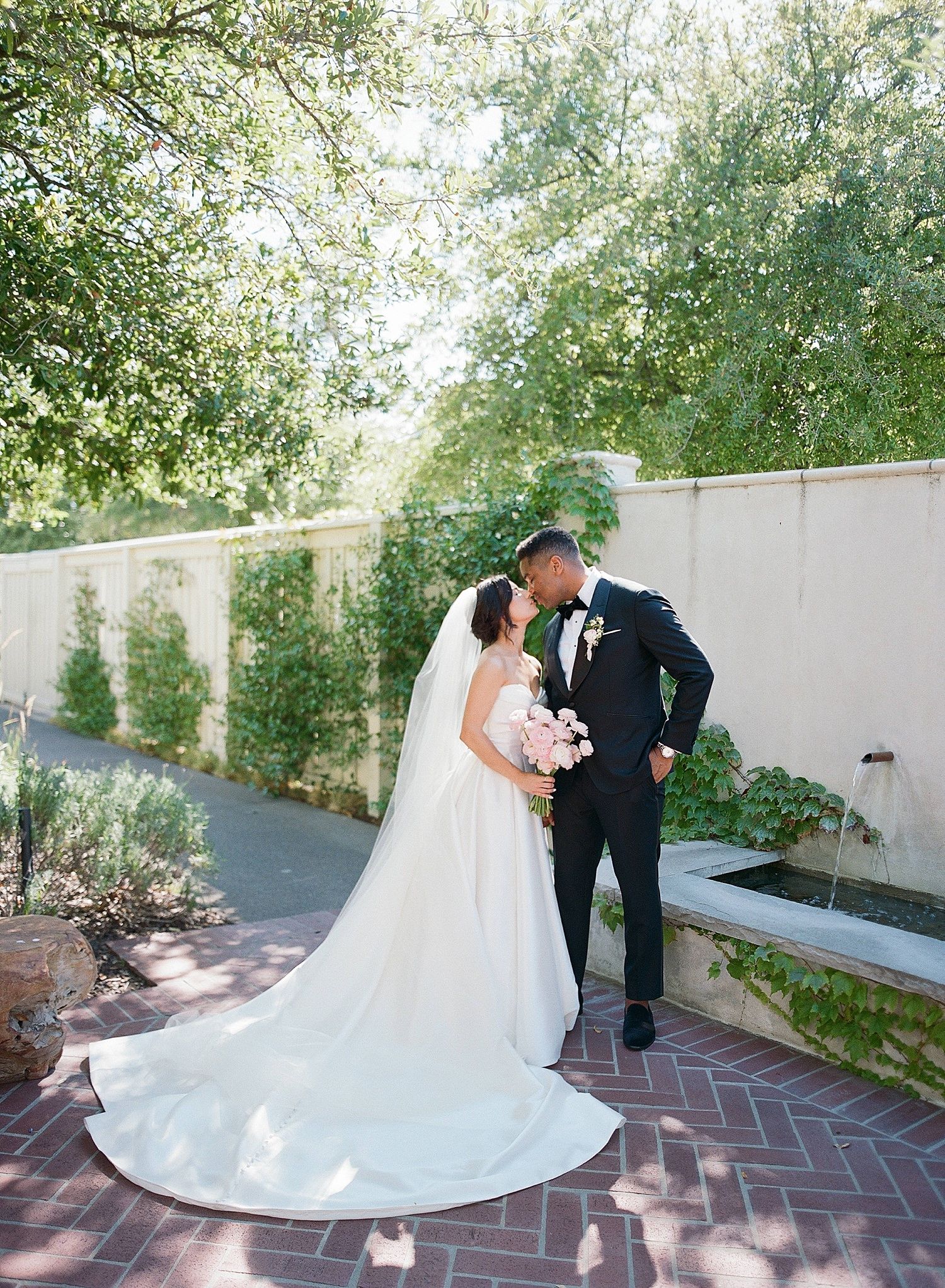 A romantic wedding photoshoot sequence captured on a brick pathway surrounded by lush greenery and white walls.