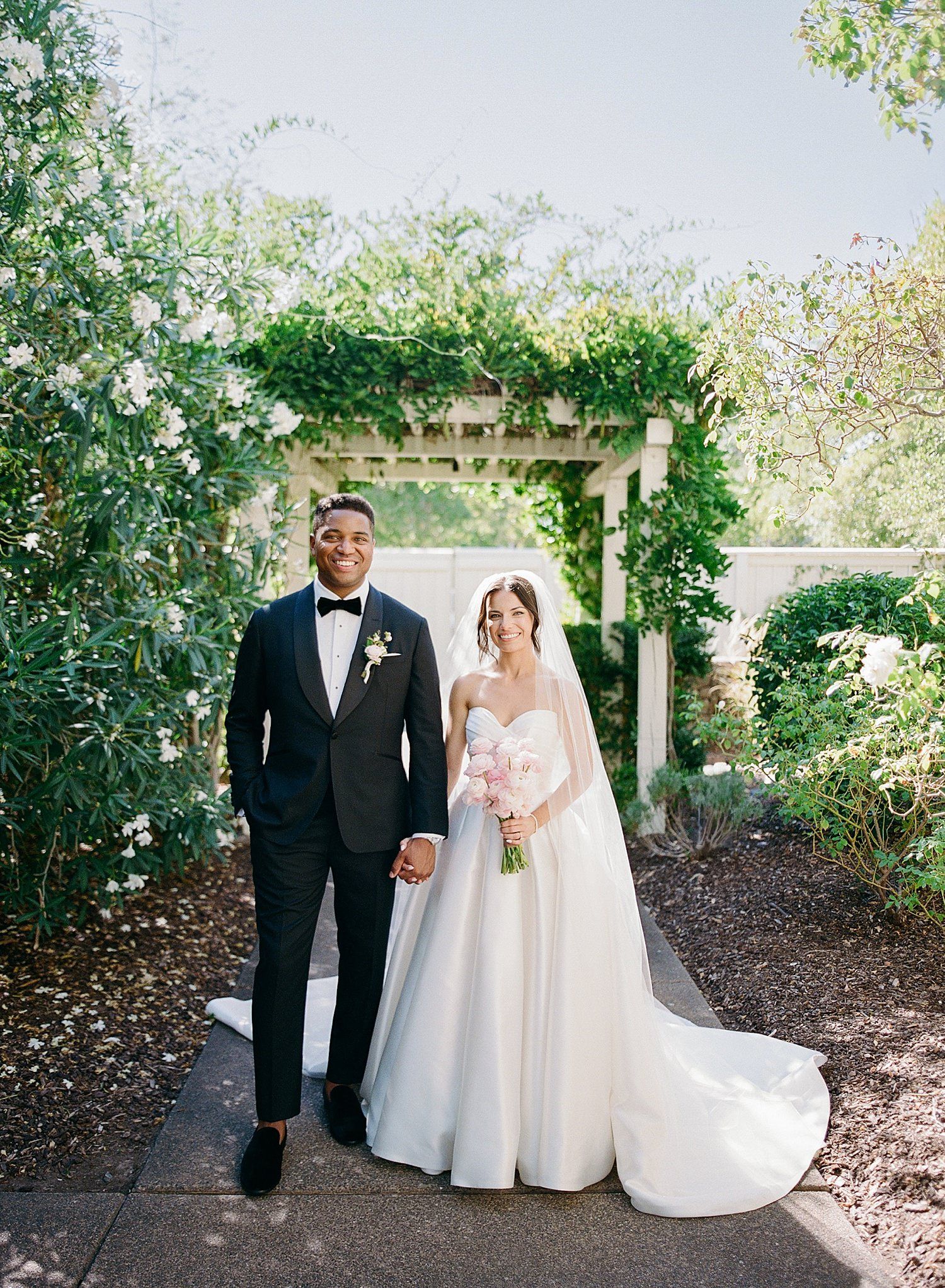 A bride and groom share a romantic moment on a garden path beneath an outdoor pergola.