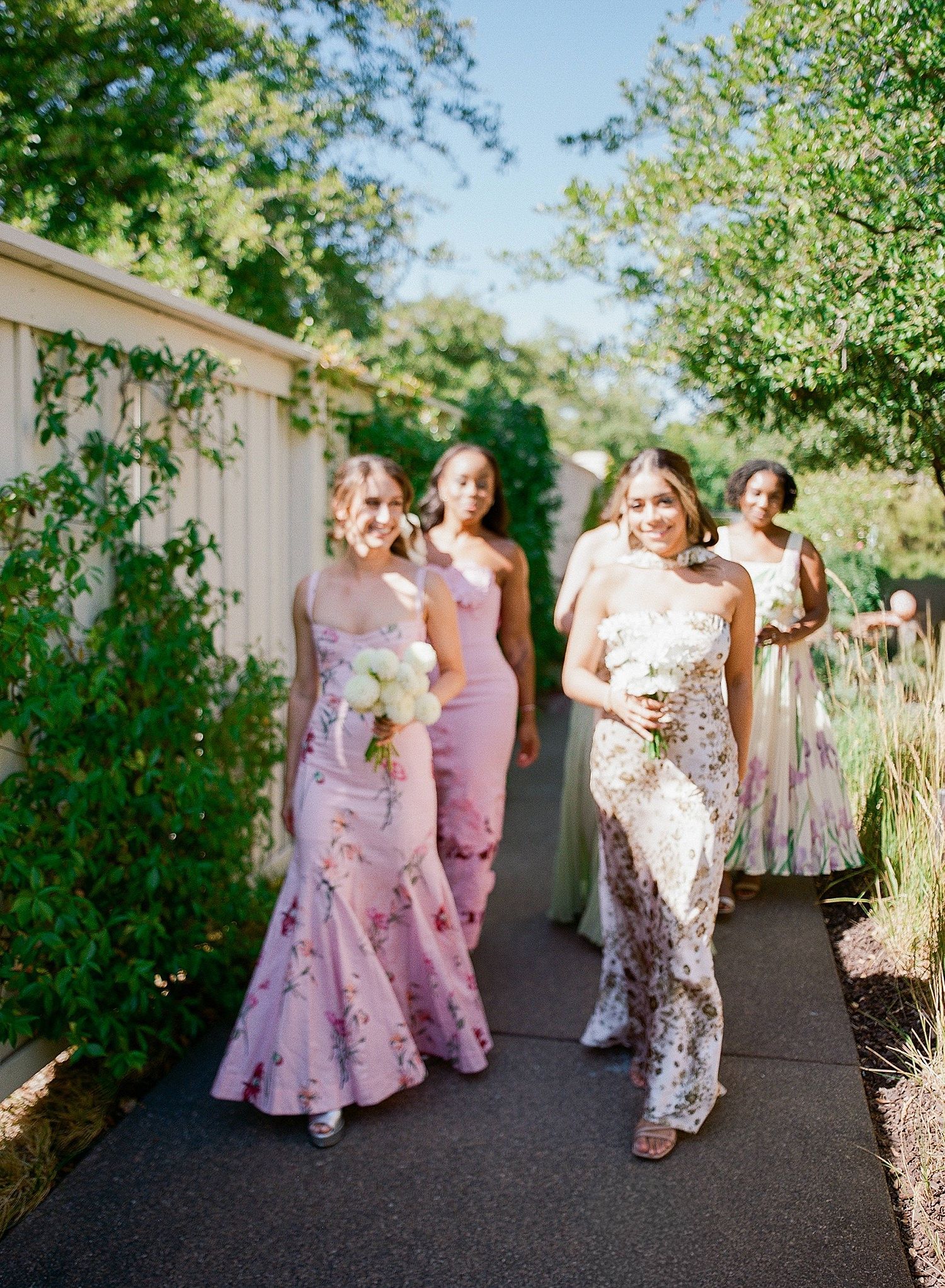 Bridesmaids in pink floral dresses walk and laugh together along a garden pathway.