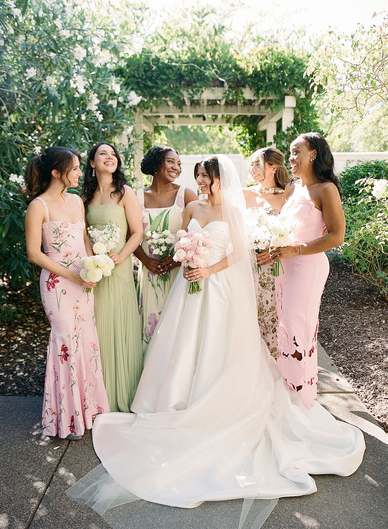 A bridal party in pink and sage green dresses poses together in a garden setting during a wedding celebration.