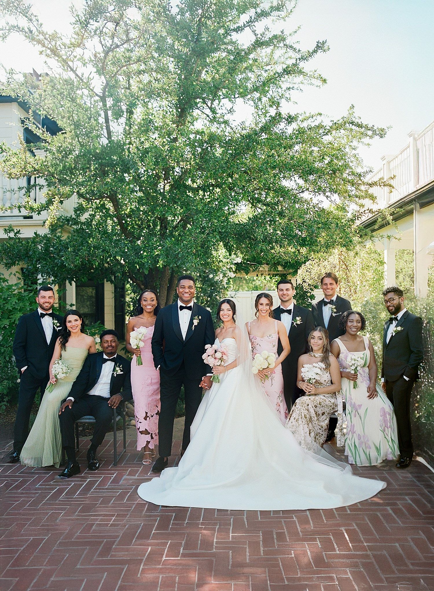 Wedding party poses together on a brick patio surrounded by lush greenery on a sunny afternoon.