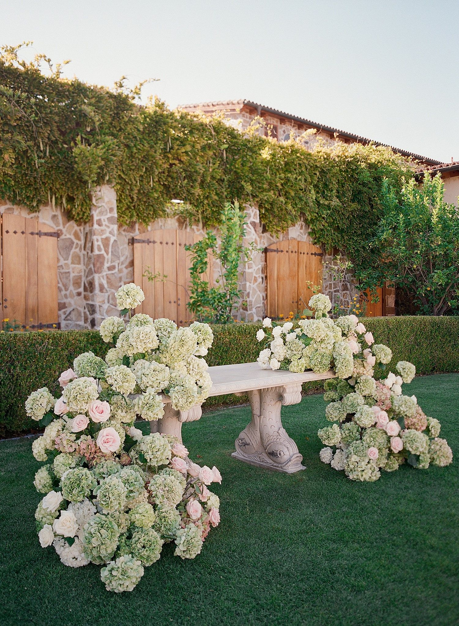 Elegant white and blush floral arrangements line a garden pathway creating a romantic outdoor ceremony setting.