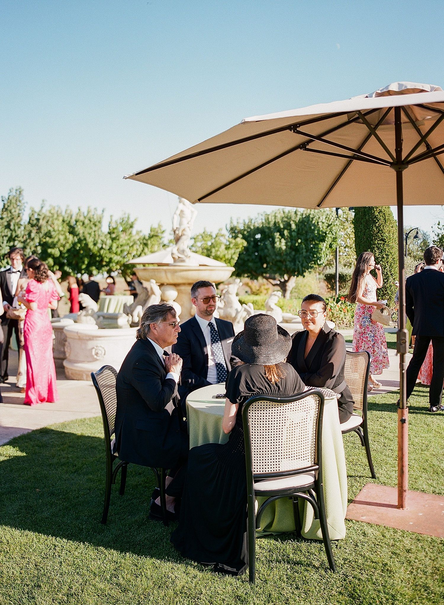 Wedding guests mingle and socialize on a grassy lawn with patio umbrellas and outdoor furniture during a daytime reception.