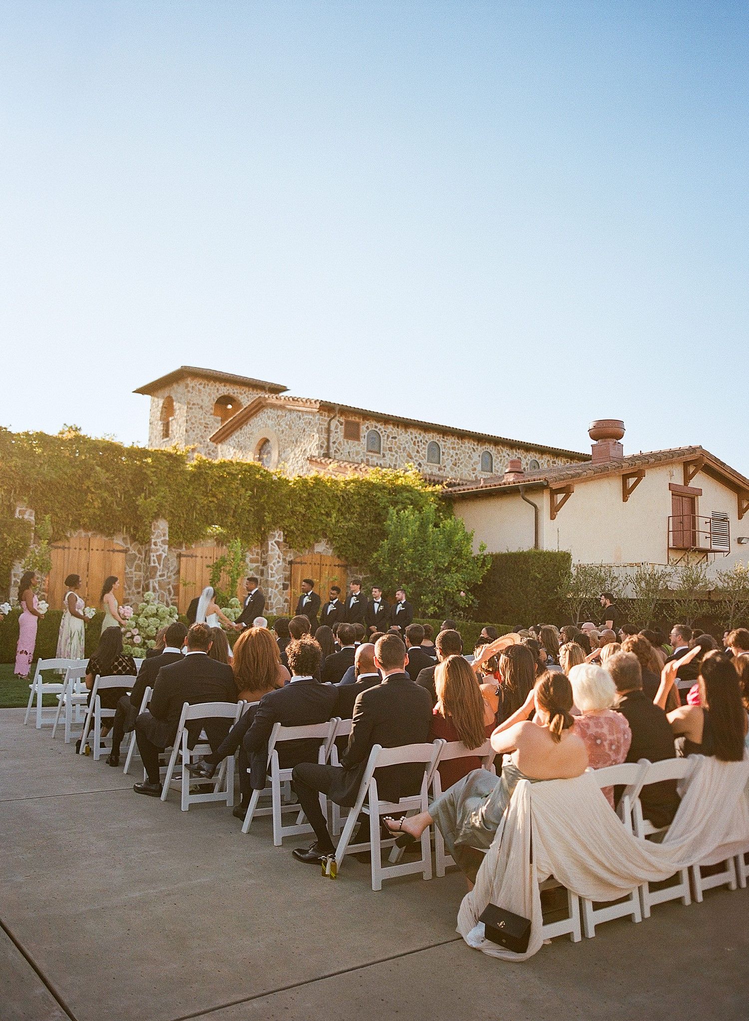 Wedding guests seated in white chairs at an outdoor ceremony venue during golden hour with Spanish-style architecture.