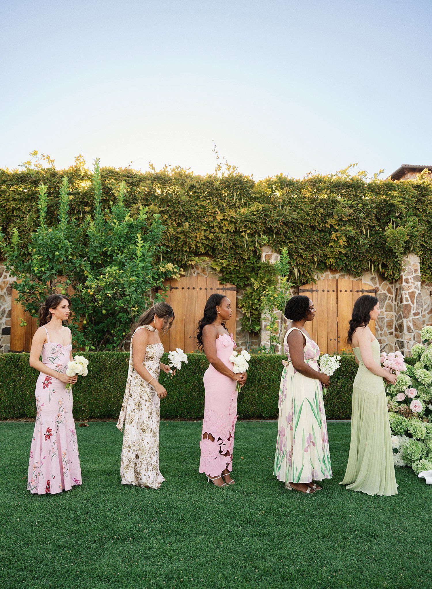 A group in pastel floral dresses stands in a line on green grass for a coordinated outdoor photo shoot.
