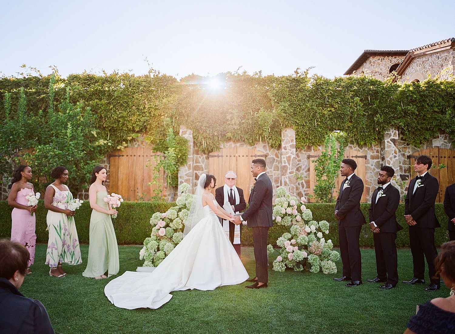 Wedding party poses together on a sunlit lawn during an outdoor garden ceremony at golden hour.