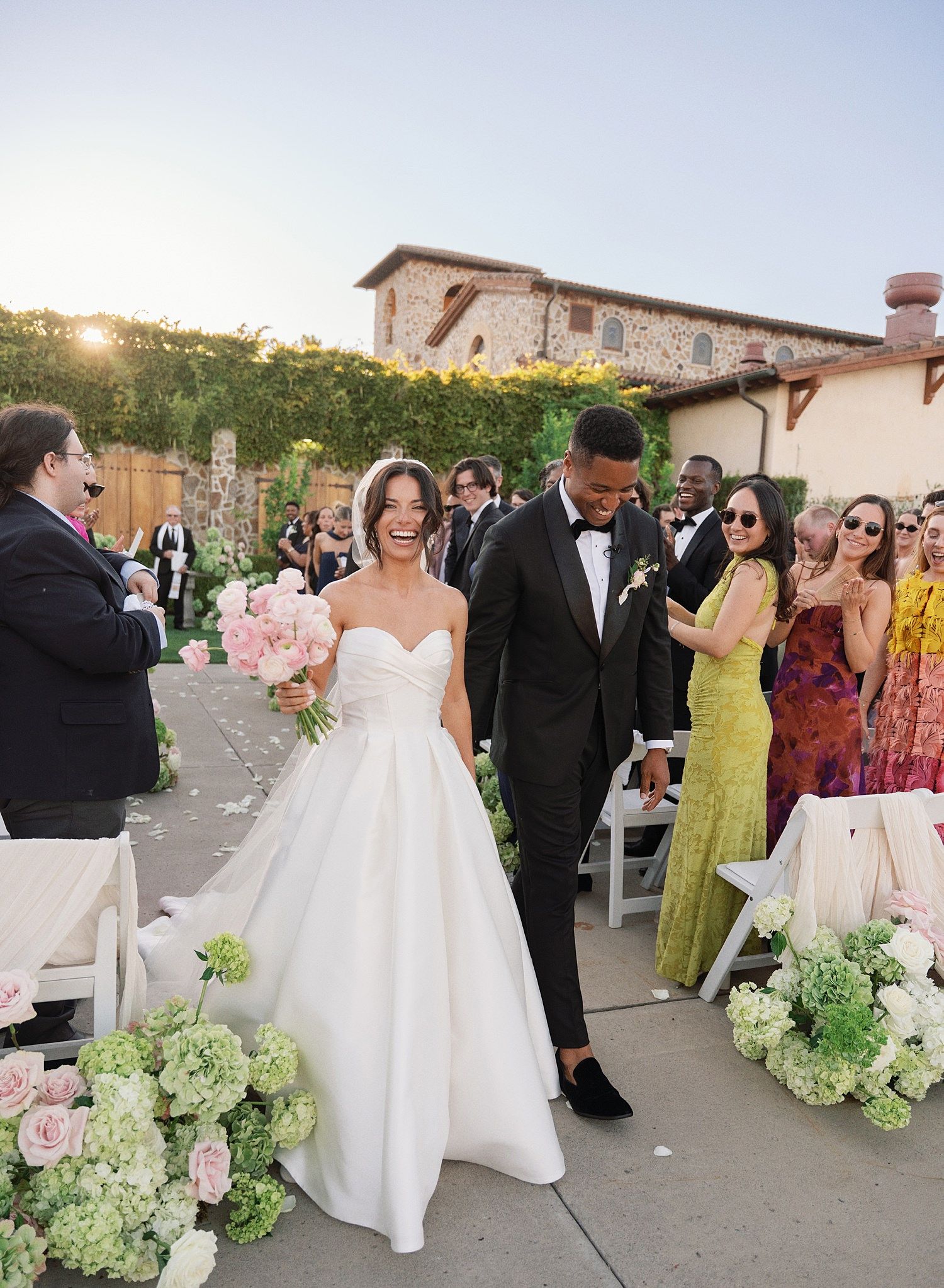 A wedding celebration with guests in formal attire and yellow dresses gathered outdoors at a scenic venue with floral arrangements.