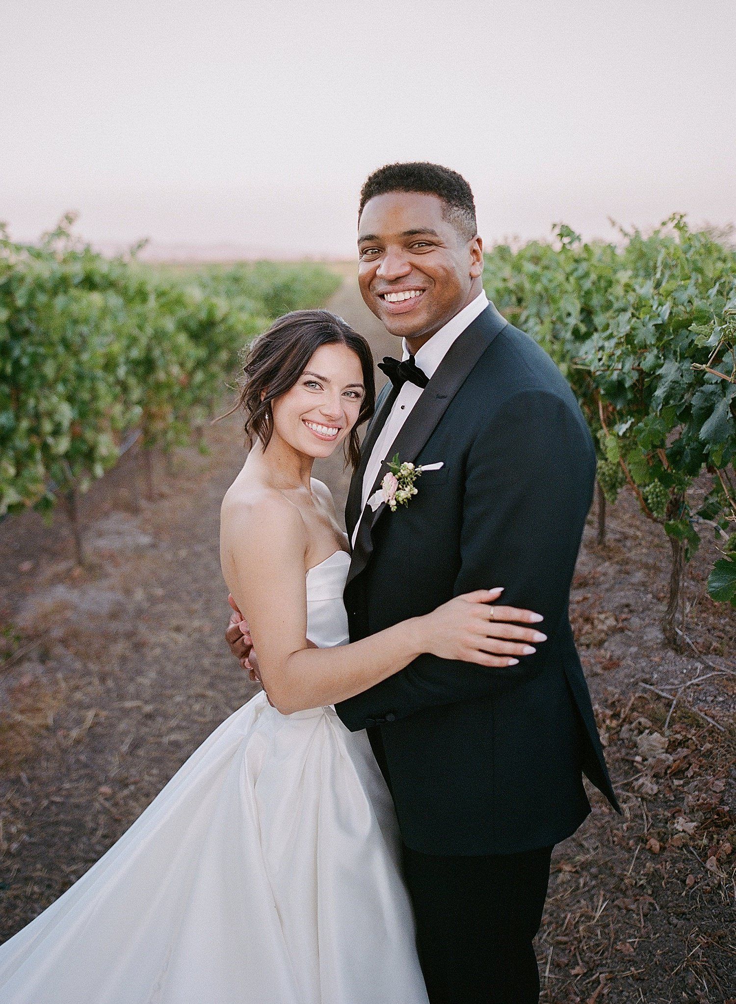 A happy couple embraces in a vineyard at sunset, the bride in white and groom in black tuxedo.