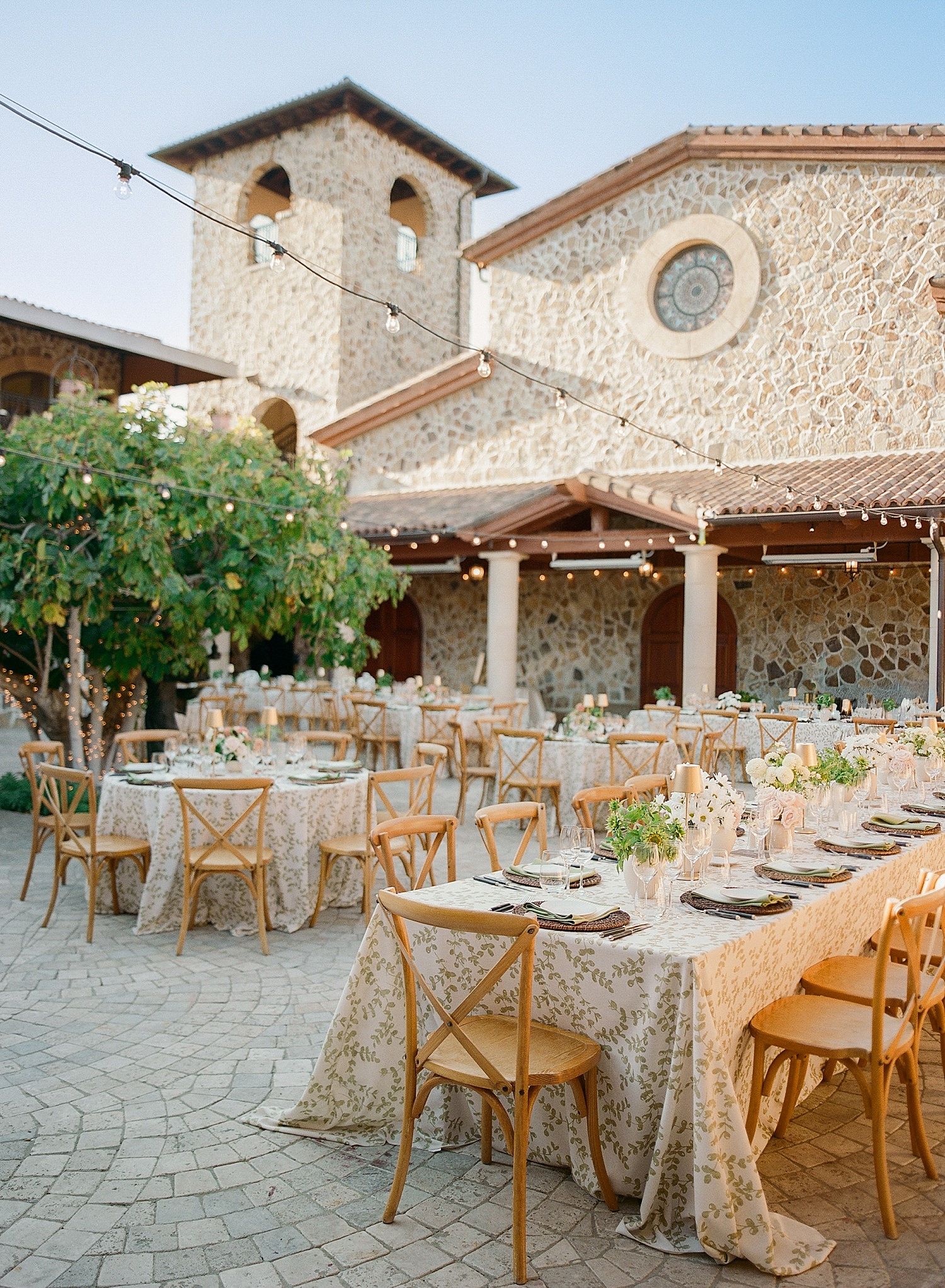 Rustic outdoor wedding reception setup with wooden chairs and tables in a Mediterranean stone courtyard.