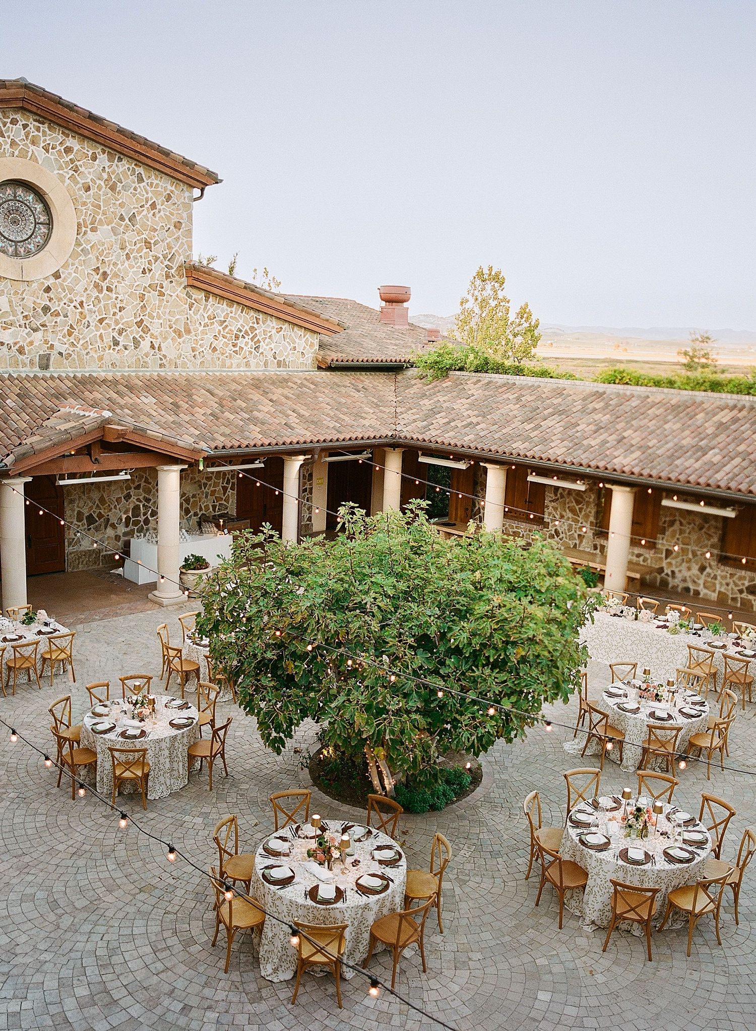 Elegant outdoor reception venue with round dining tables and chairs arranged on a stone patio surrounded by Mediterranean architecture.