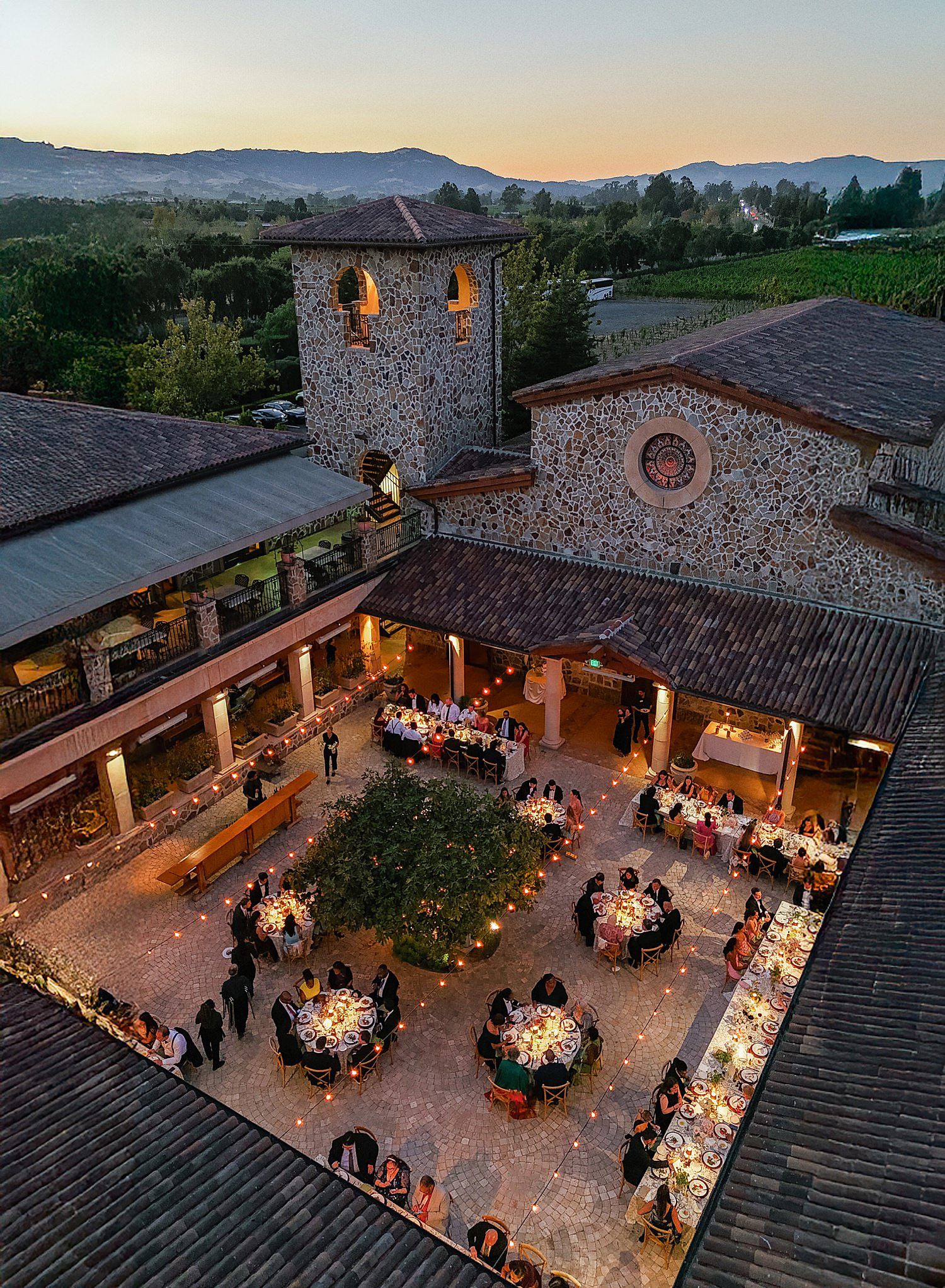 Outdoor wedding reception venue at sunset with long dining tables and string lights illuminating a rustic stone courtyard.