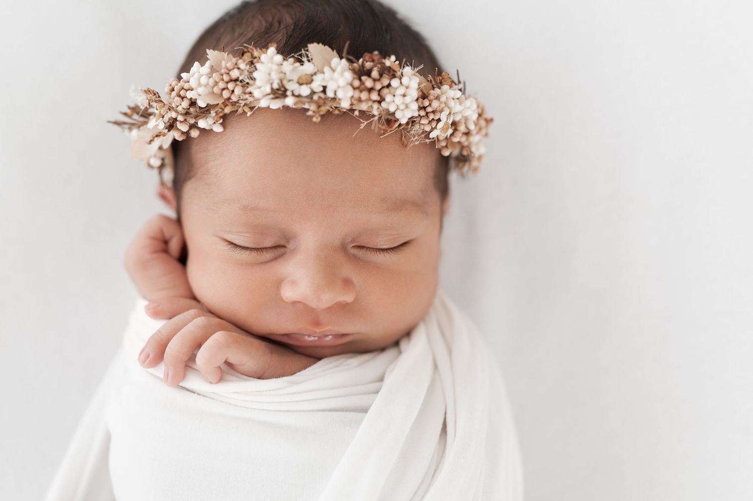 Sleeping newborn wearing delicate floral crown and white wrap in minimalist photography setup.