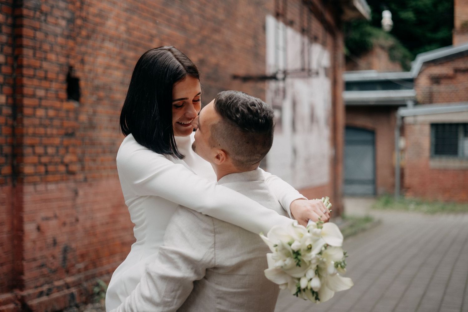 A couple in white attire shares an intimate moment along a brick wall with white floral bouquet.