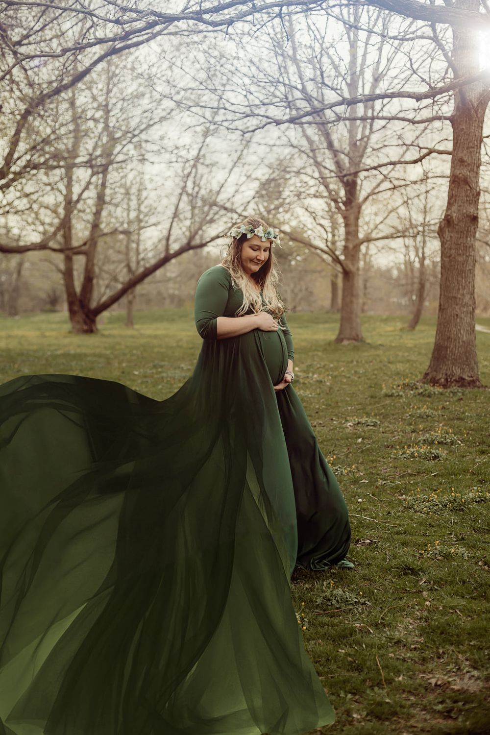 Maternity photo of woman in flowing green dress standing among bare trees.