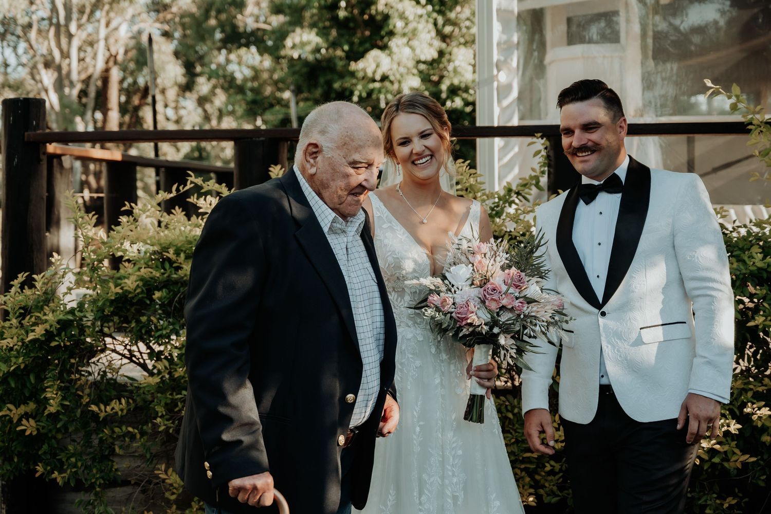 Wedding party in formal attire stands together outdoors in a garden setting during a celebration.