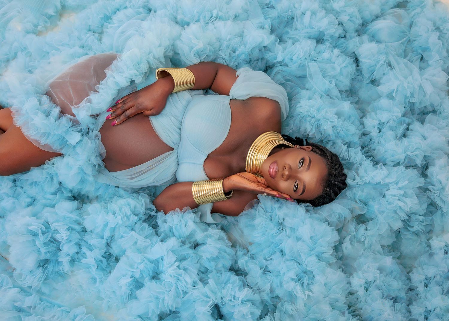 Pregnant woman in light blue outfit reclines on fluffy blue tulle cloud with gold accessories.