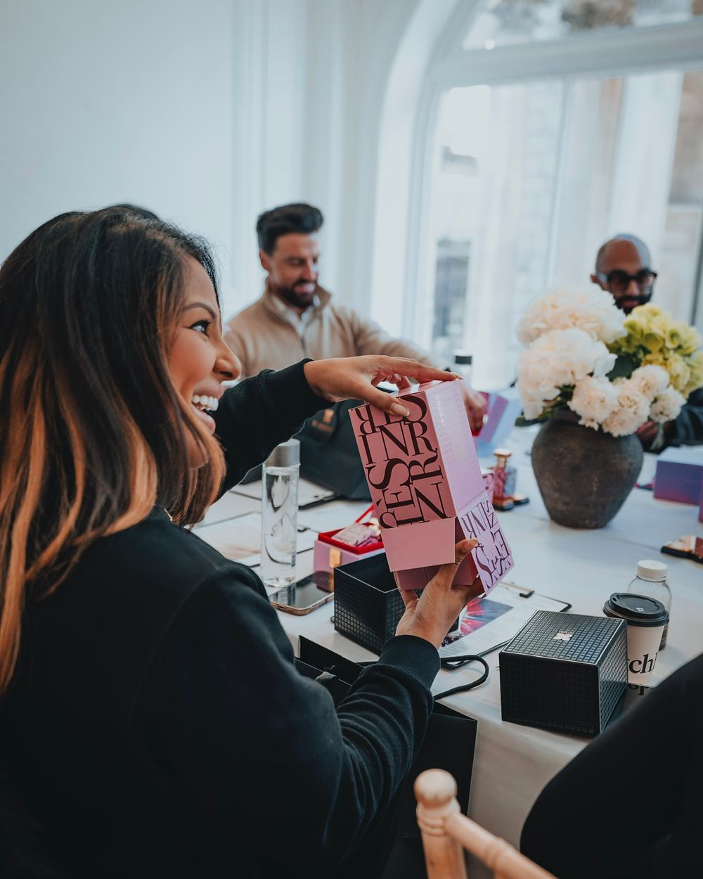 People enjoying coffee and treats at a bright cafe table with pink cards and floral decorations.