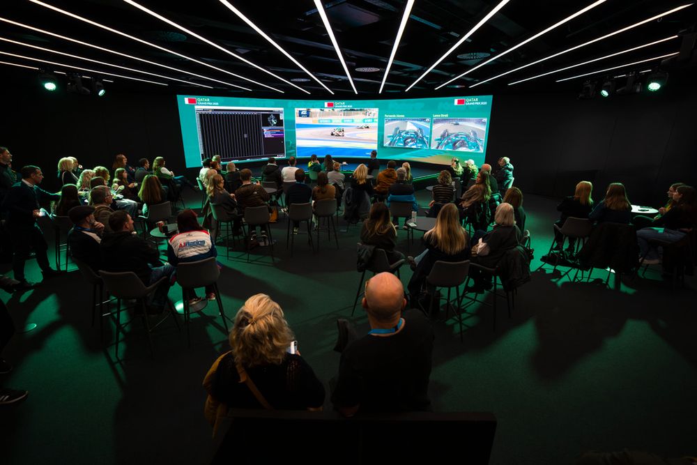 A dark auditorium filled with people watching a presentation on large screens with teal lighting effects.